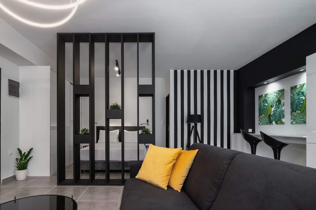 Un salon moderne avec un canapé noir, un coussin jaune, une table basse en verre, un lampadaire noir et un mur orné de rayures verticales noires et blanches.