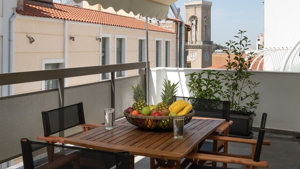 Ein Holztisch mit Obstschale und Gläsern auf einem Balkon mit Blick auf ein Gebäude und einen Glockenturm.