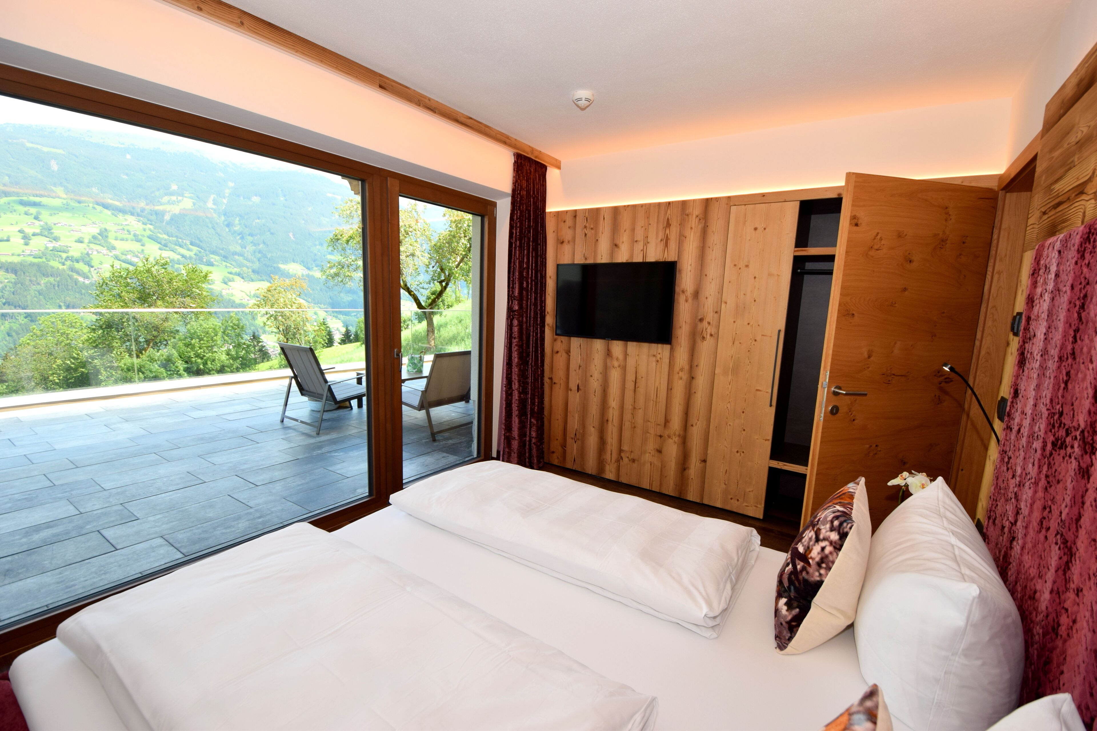 A bedroom with a large bed, a TV mounted on the wall, and a sliding door leading to a balcony with a view of the countryside.