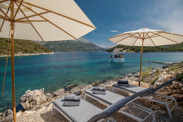 A coastal view with lounge chairs and umbrellas, a boat in the water, and rocky terrain. - Luxury Villa Korcula Supreme (Korcula)