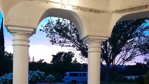A covered outdoor seating area with a sofa and cushions, a view of a vehicle in the background, and a sunset sky. - Private Suite (Paso Robles)