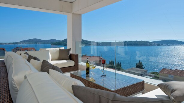 A balcony with a view of a body of water, a wicker sofa, and a glass table with a bottle and glass. - Luxury Villa Carpe Diem Paradise (Rogoznica)