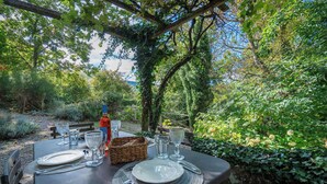 Ein Essplatz im Freien mit Geschirr, Besteck und Gläsern, umgeben von üppigem Grün und einer Pergola.