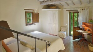 Ein Schlafzimmer mit einem Bett, einem kleinen Fenster, einem Kleiderschrank, einem Stuhl, einem Schreibtisch mit Lampe und einer Tür, die nach draußen führt.