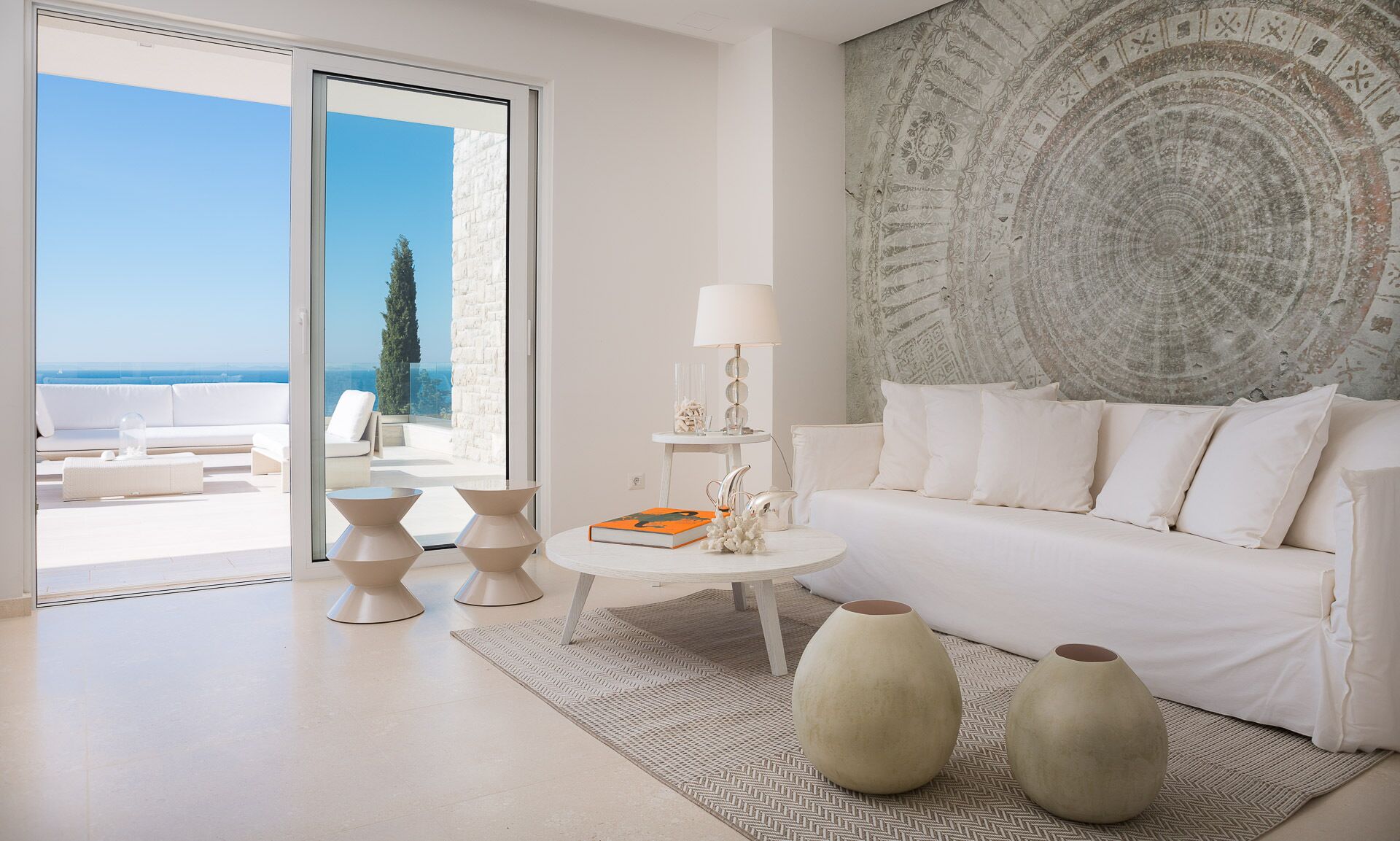 Un salon moderne avec un canapé blanc, une table basse ronde et deux vases. Une grande tapisserie orne le mur, et un balcon avec vue sur la mer est visible à travers les portes vitrées.