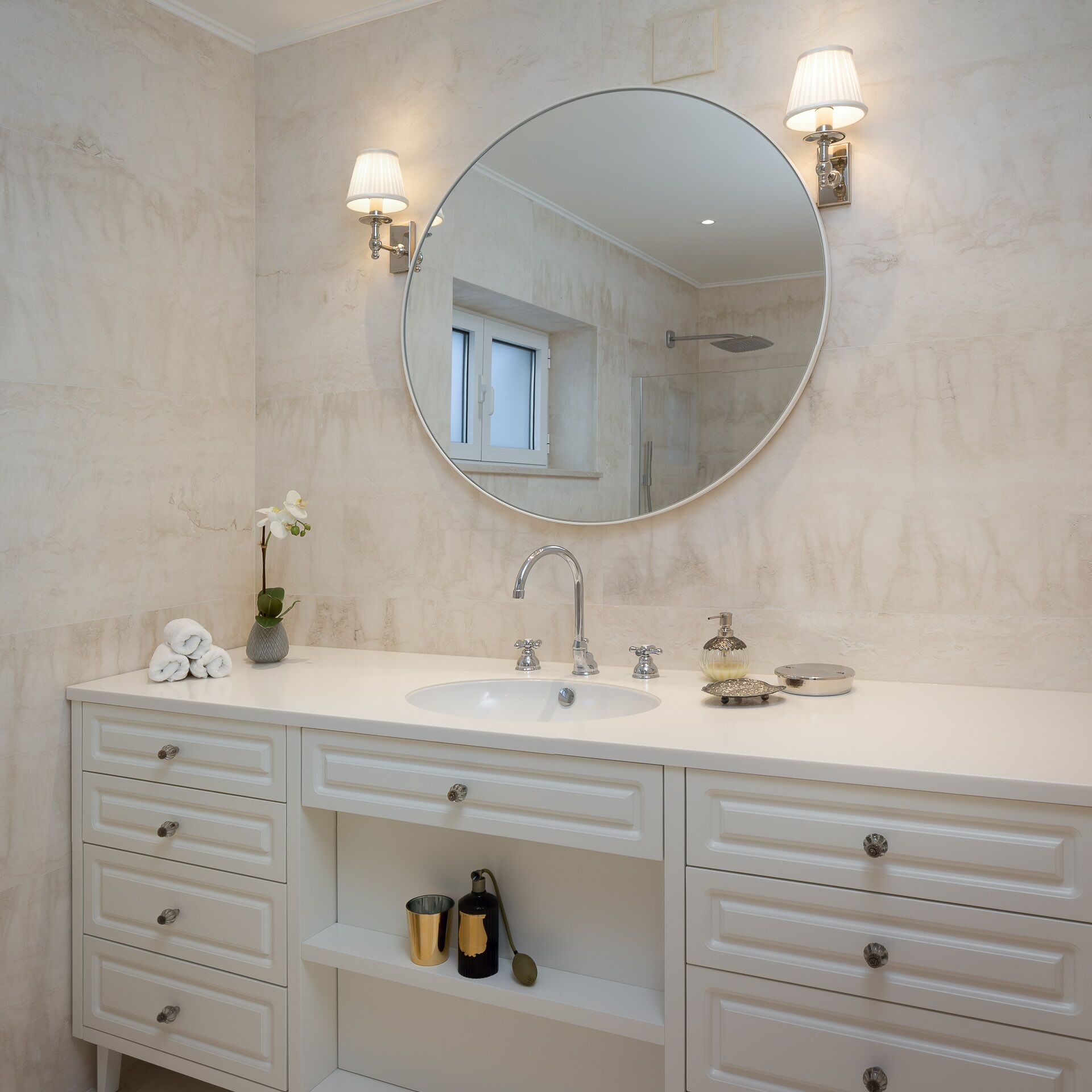 A bathroom with a round mirror, a sink, and two wall-mounted lights. There are towels on the counter and a small shelf with bottles.