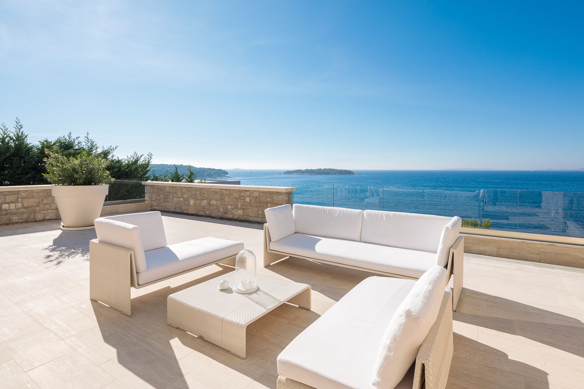 Una terrazza con mobili da esterno bianchi, una pianta in vaso e vista sul mare.