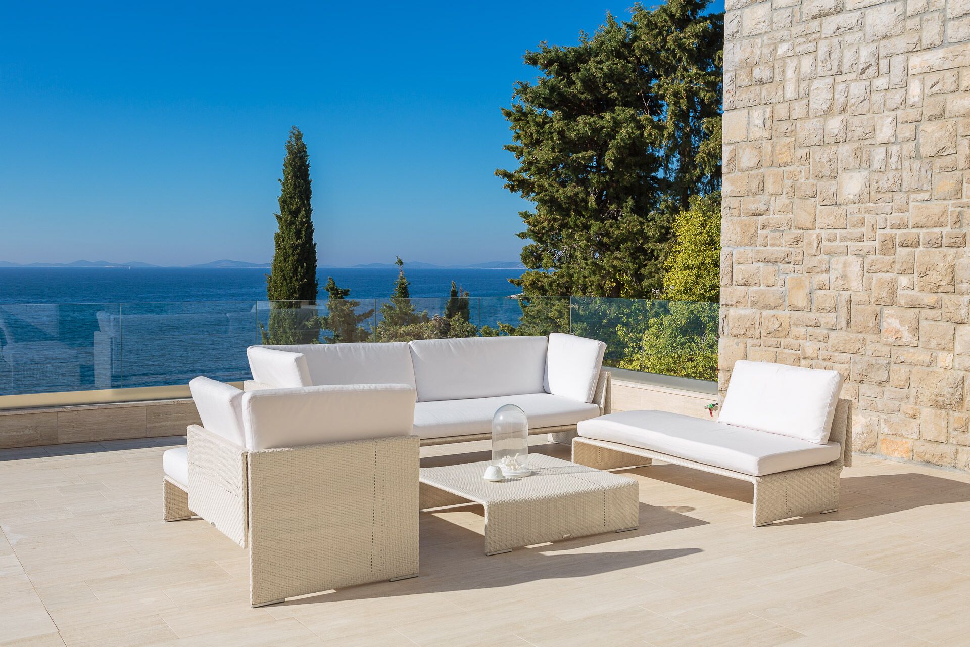 A balcony with a white wicker sofa set and a glass coffee table, overlooking a scenic view of the sea and mountains.