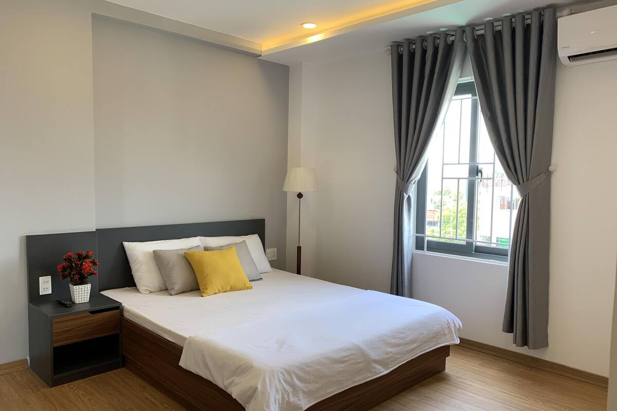 A modern hotel room with a large bed, a nightstand, a lamp, and a window with curtains.
