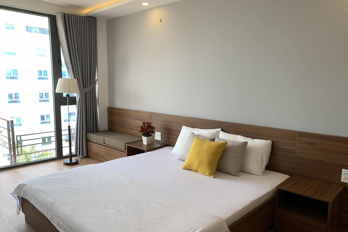 A hotel room with a large bed, a bedside table, a lamp, and a view of the city through the window.