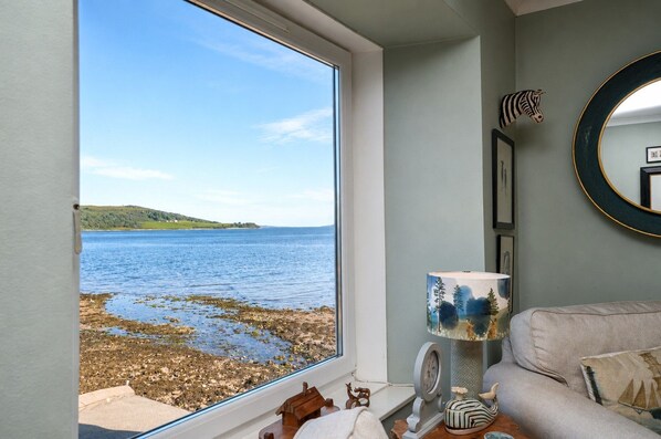 A room with a large window overlooking a rocky shoreline and calm sea, a round mirror on the wall, and a sofa with cushions.