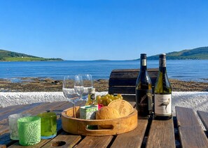 A wooden table with a bottle of wine, glasses, a bowl of grapes, and a cheese wheel by a lakeside.