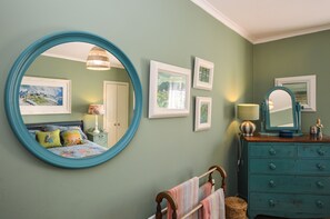 A bedroom with a large round mirror, a bed with colorful bedding, a dresser with drawers, and several framed pictures on the wall.