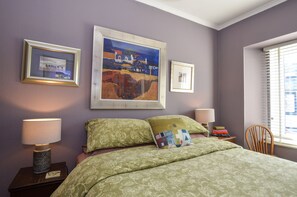 A bedroom with a bed, two bedside lamps, a window with blinds, and three framed artworks on the wall.
