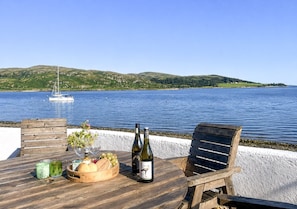 A wooden table with a bottle of wine, glasses, and a fruit bowl, overlooking a calm body of water with a sailboat in the distance.
