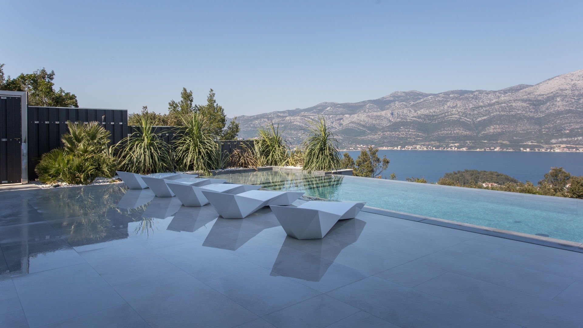 An infinity pool with white geometric loungers, surrounded by tropical plants, with a mountainous backdrop and a view of the sea.