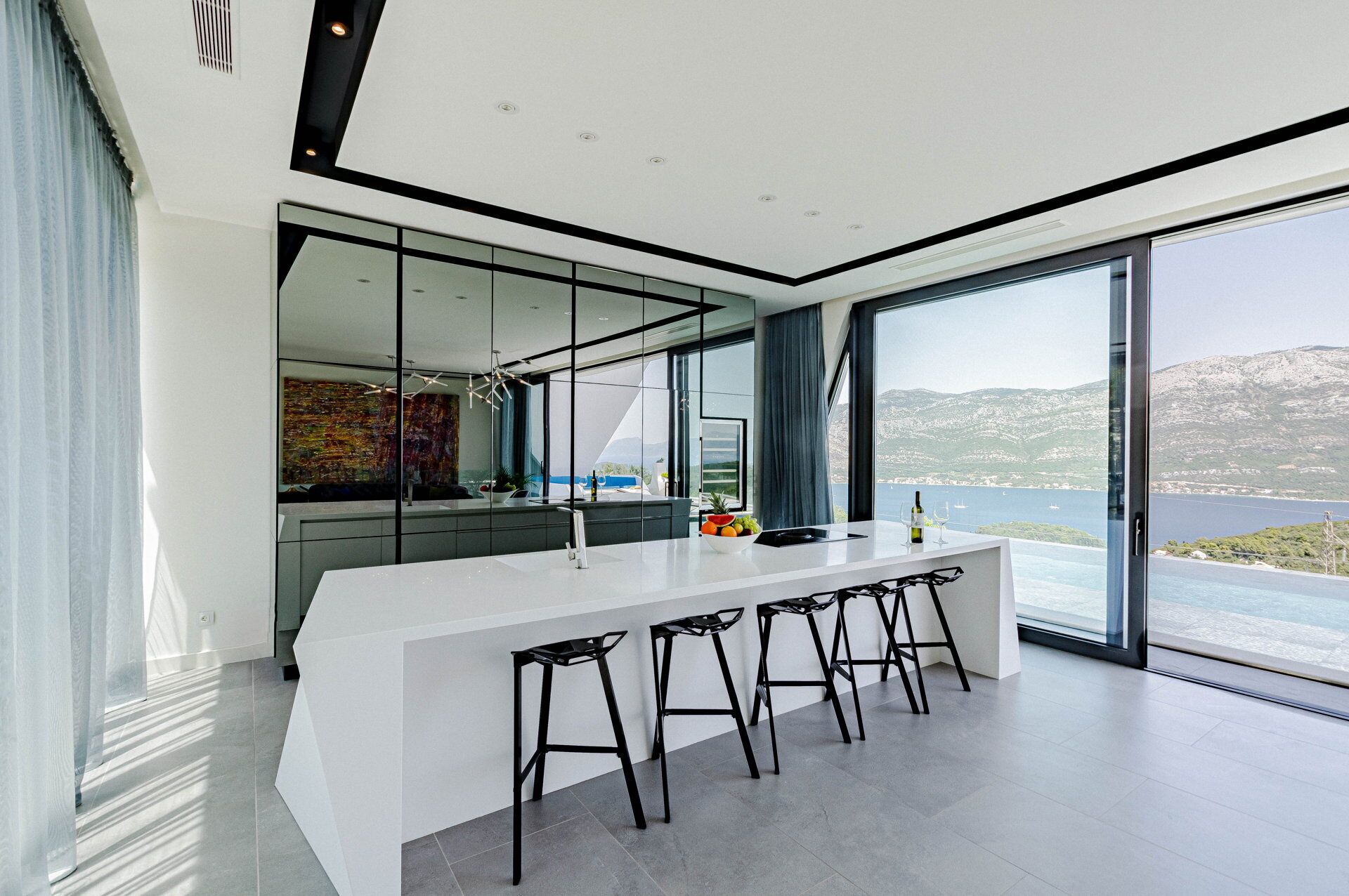 A modern kitchen with a large island, black bar stools, and a view of the sea.