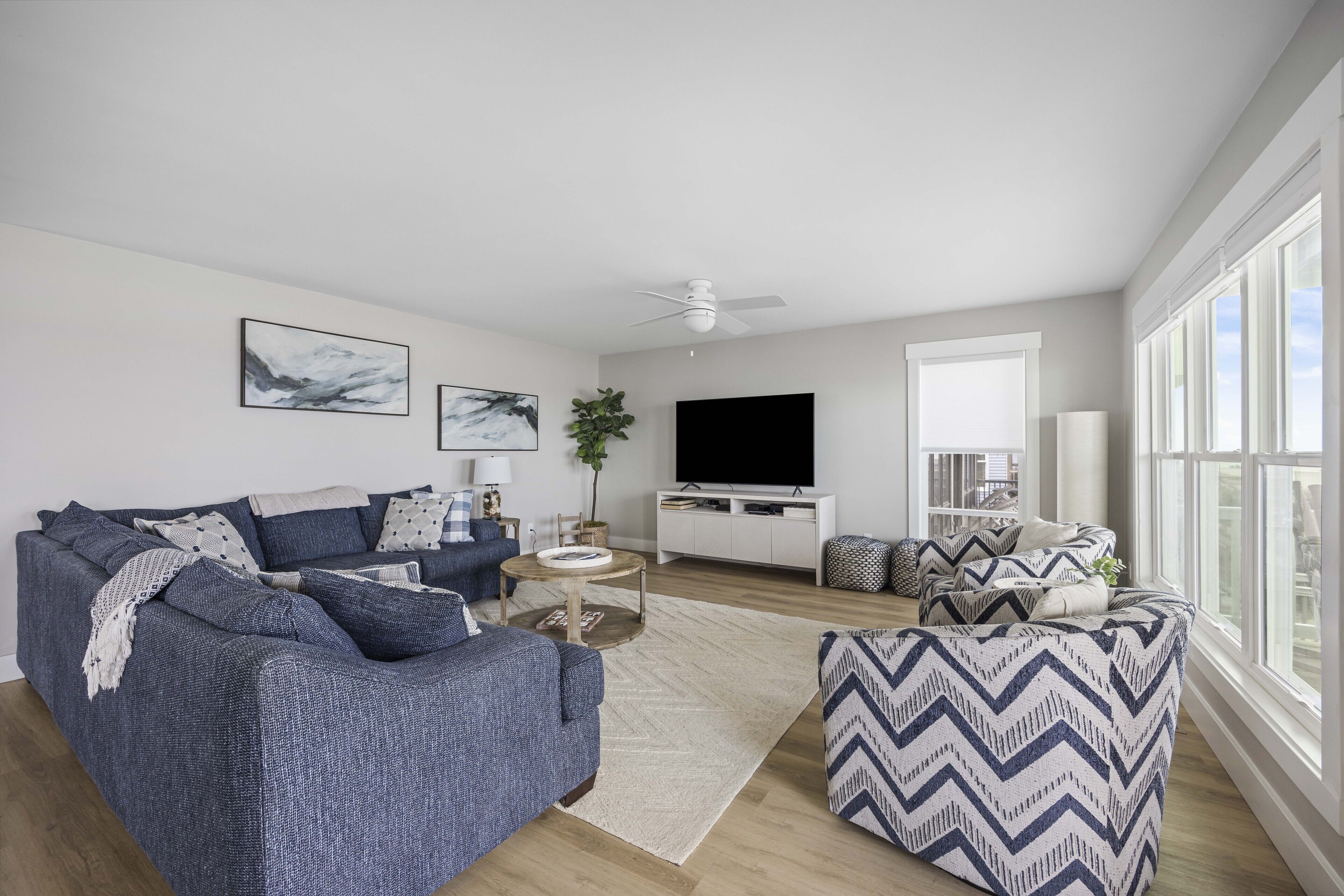 A modern living room with a grey sofa, a patterned ottoman, a flat-screen TV, and a large window.
