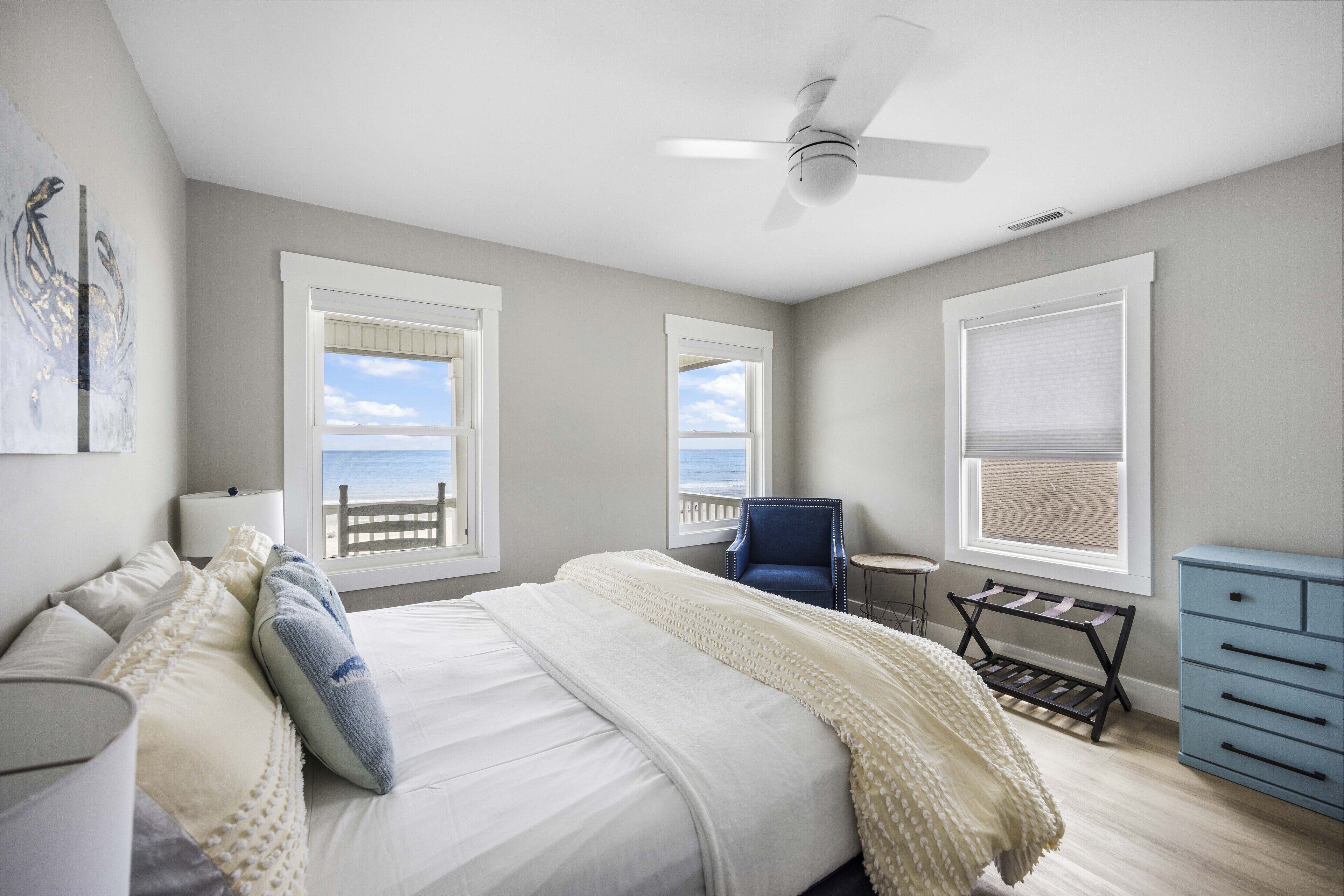 A bedroom with a bed, a chair, a small table, a ceiling fan, and a view of the ocean through windows.