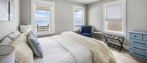 A bedroom with a bed, a chair, a small table, a ceiling fan, and a view of the ocean through windows.