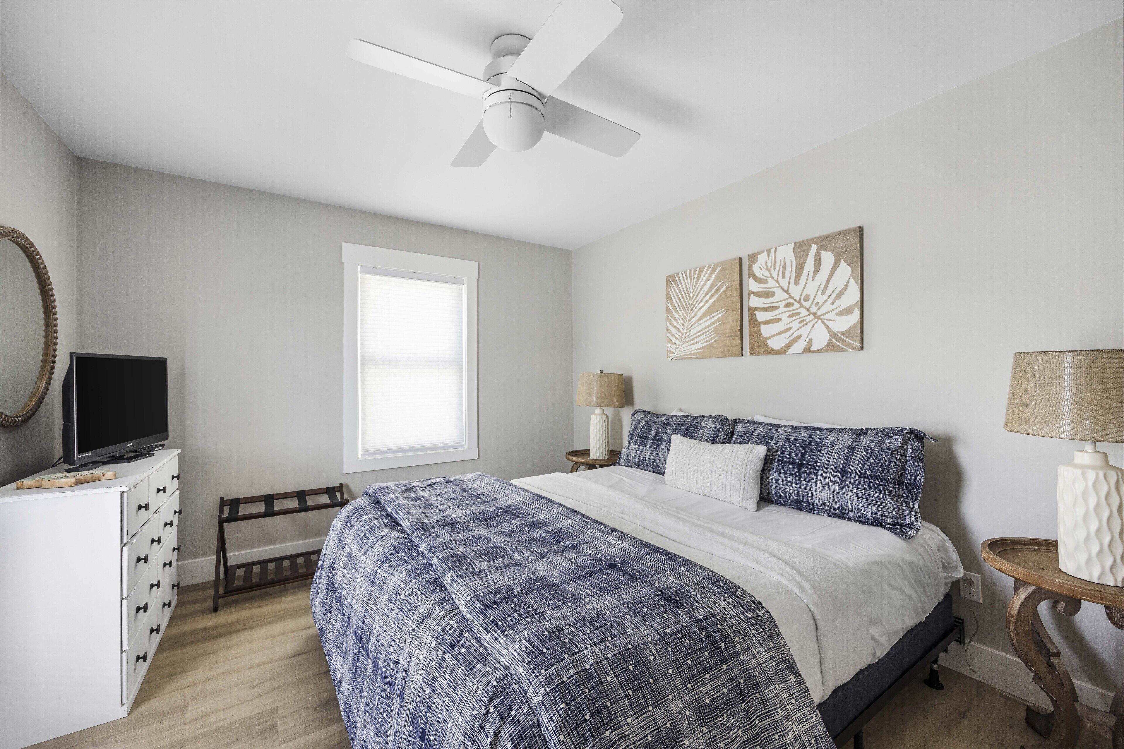 A bedroom with a bed, a ceiling fan, a TV on a dresser, a window, and artwork on the wall.
