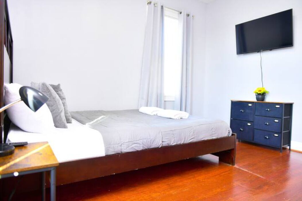 A bedroom with a bed, a TV mounted on the wall, a dresser, and a window with curtains.