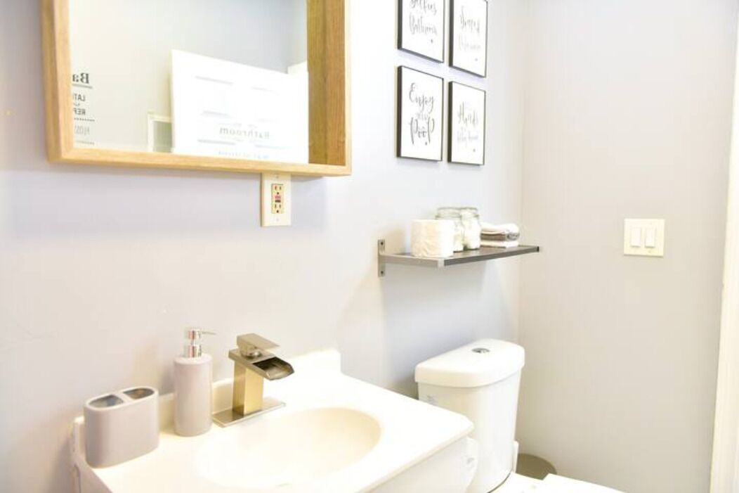 A bathroom with a white sink, toilet, and a mirror above the sink. There are framed pictures on the wall and a shelf with toiletries.
