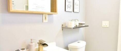 A bathroom with a white sink, toilet, and a mirror above the sink. There are framed pictures on the wall and a shelf with toiletries.
