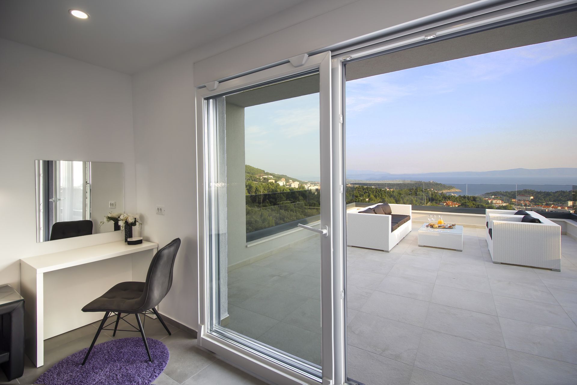 A modern balcony with a city view, a white desk with a chair, and a large sliding glass door.