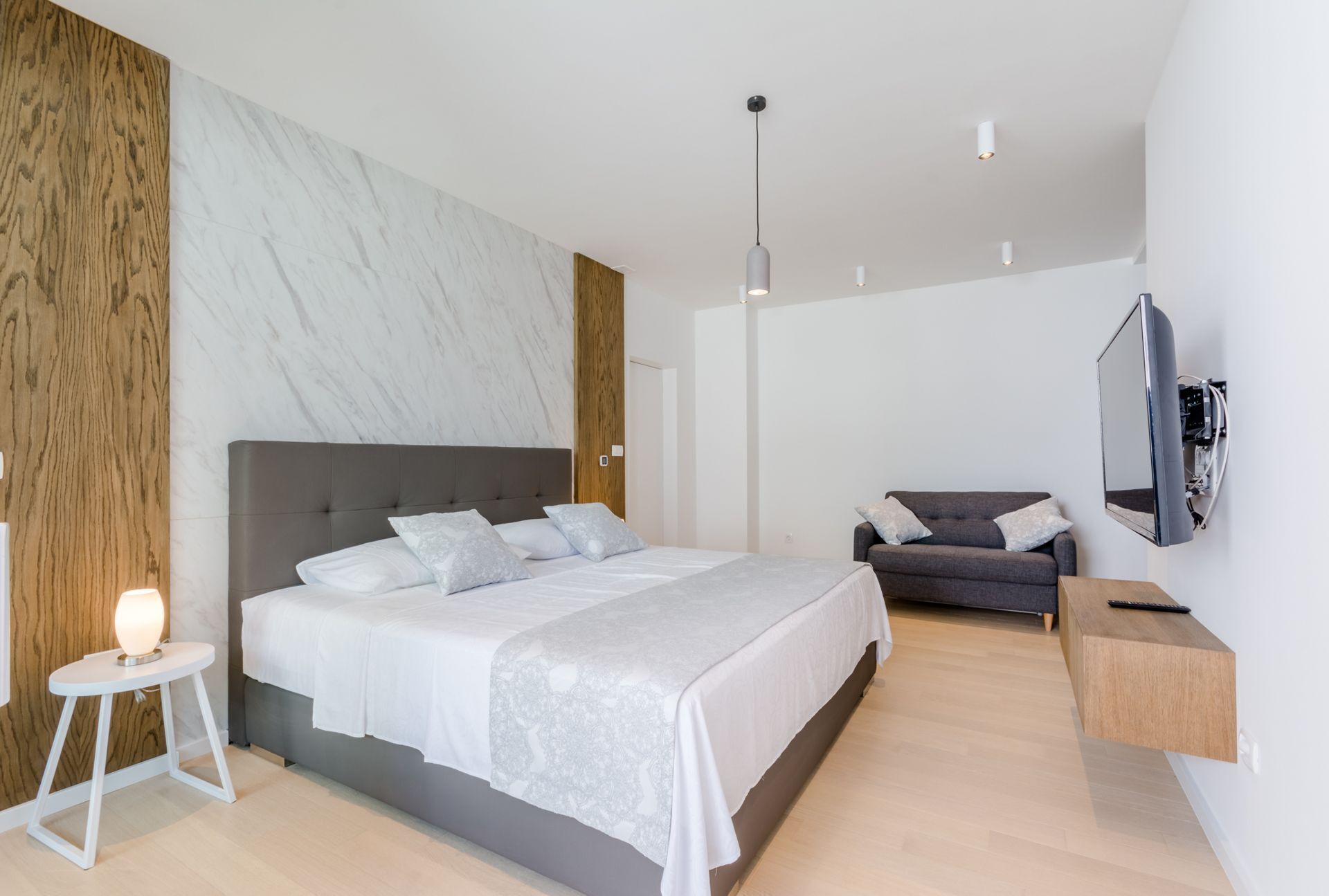 Un dormitorio moderno con una cama grande, un sofá gris, un televisor montado en la pared y una pared de acento de madera.