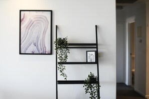 Un couloir orné d’un tableau abstrait encadré, d’une étagère en métal noir agrémentée de plantes vertes et d’une porte visible au fond.