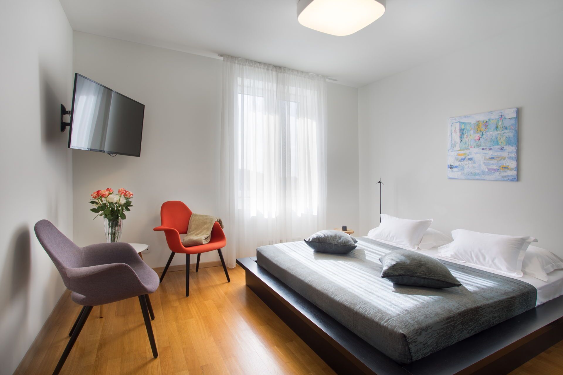 Een moderne hotelkamer met een bed, een stoel, een klein tafeltje, een televisie en een schilderij aan de muur.