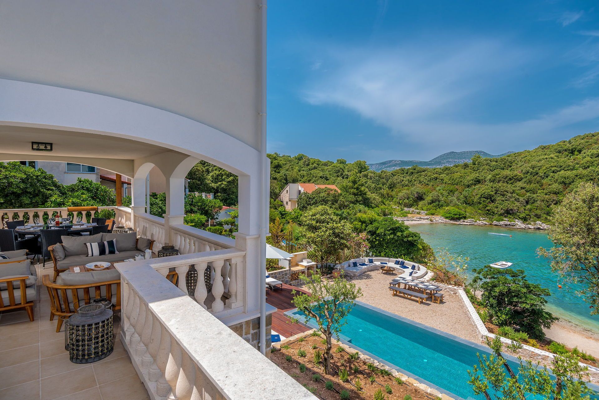 Un balcon avec coin salon extérieur, une piscine et une vue sur une zone côtière verdoyante, sous un ciel dégagé.