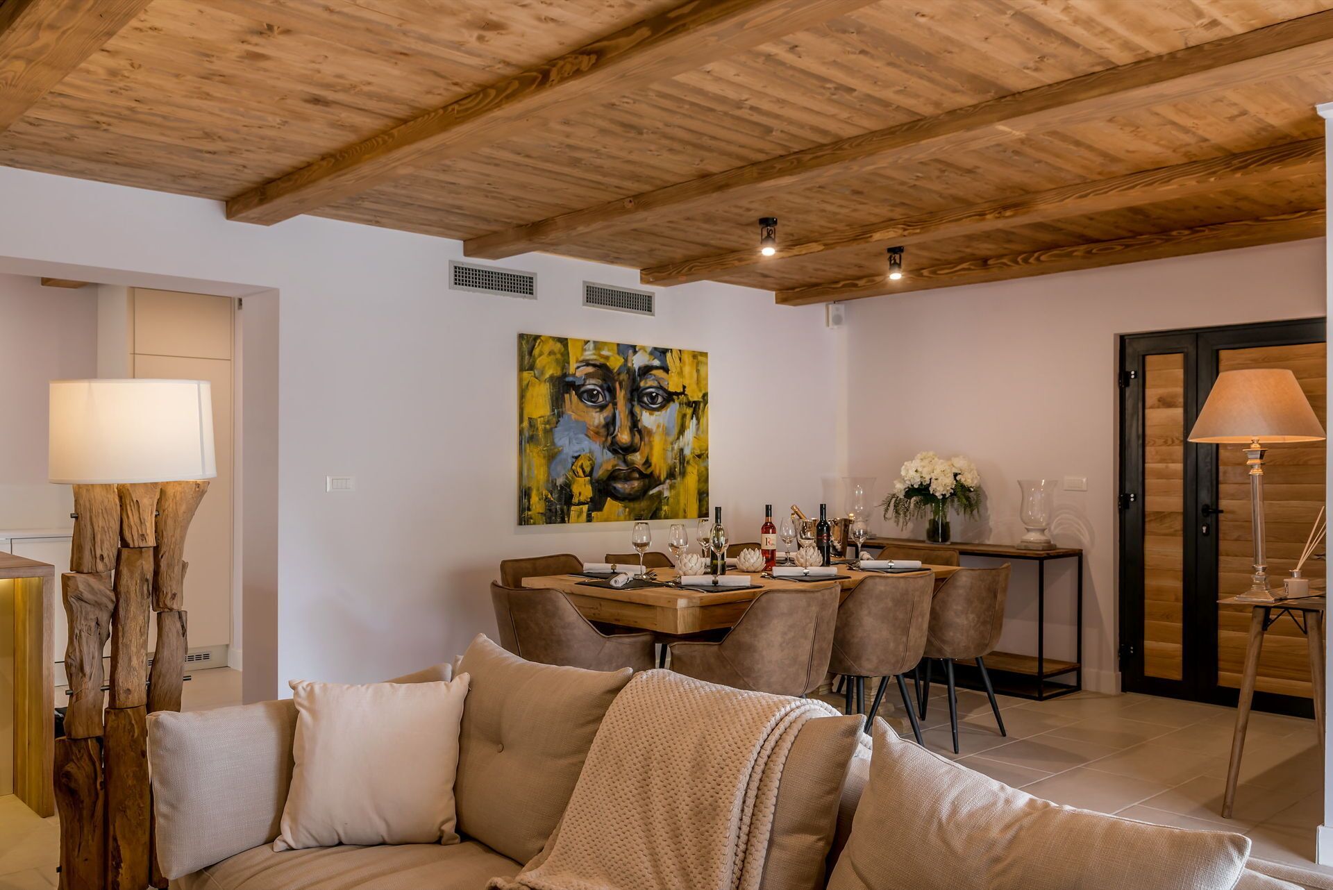 A cozy living room with a wooden ceiling, a dining area with a table set for four, and a large abstract painting on the wall.