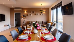 Une salle à manger moderne avec une longue table dressée pour six personnes, un canapé en arrière-plan et une grande fenêtre offrant une vue dégagée.