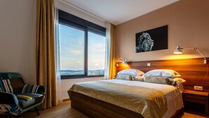 Une chambre d’hôtel avec un grand lit, une chaise, une fenêtre avec des rideaux, une tête de lit et un tableau au mur.