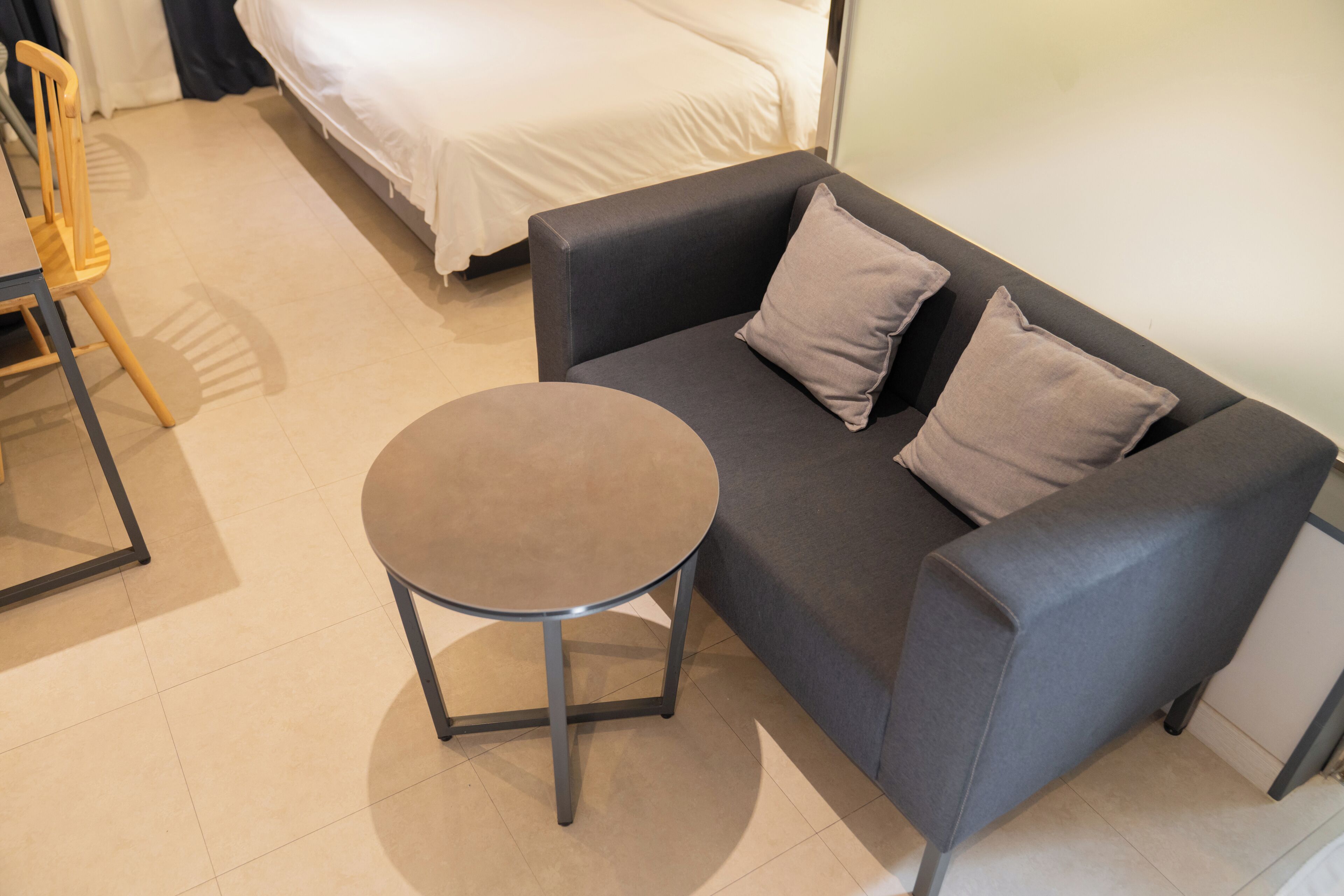 Una habitación con un sofá gris, dos almohadas, una mesita redonda pequeña, una silla de madera y una cama con ropa blanca.