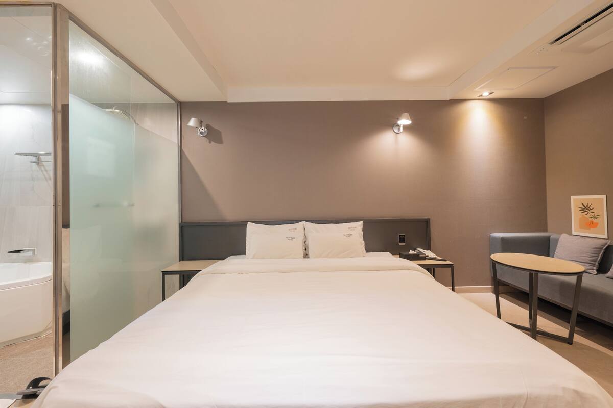 A hotel room with a bed, a small table, a sofa, a glass shower enclosure, and a wall-mounted light fixture.