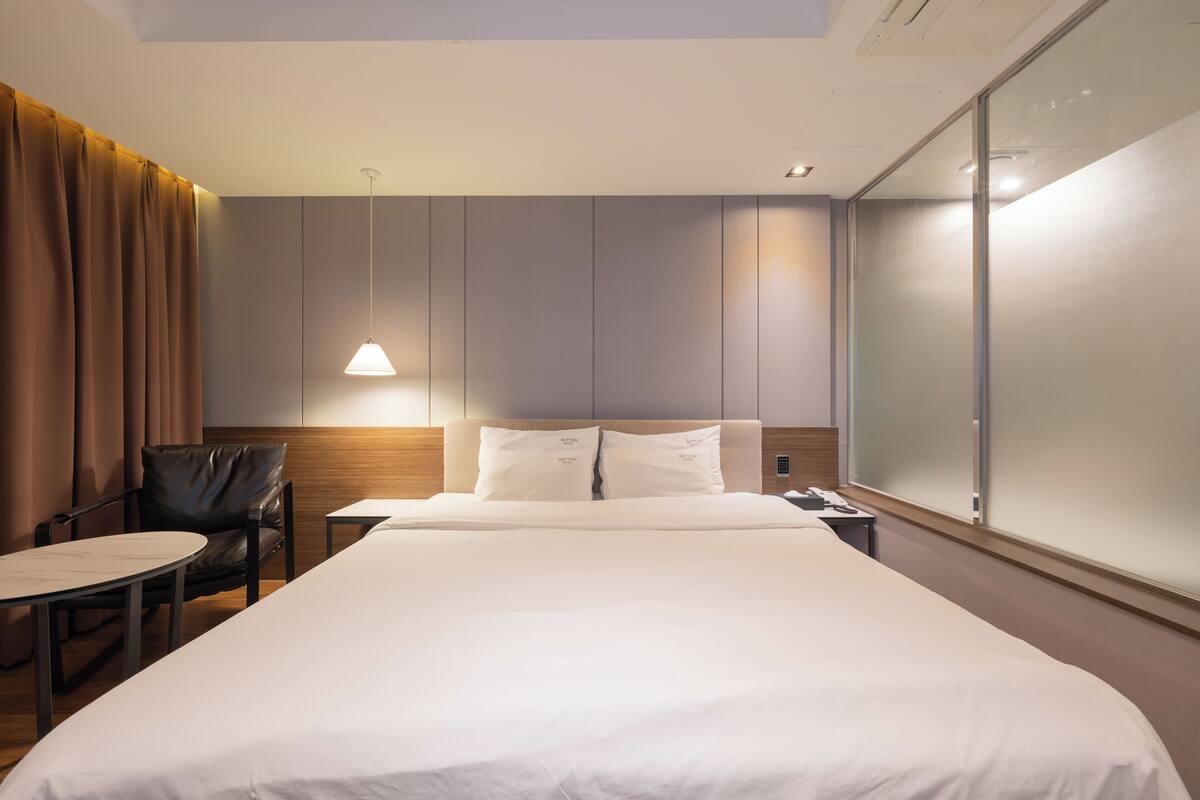 A hotel room with a large bed, a chair, a small table, and a wall-mounted phone.