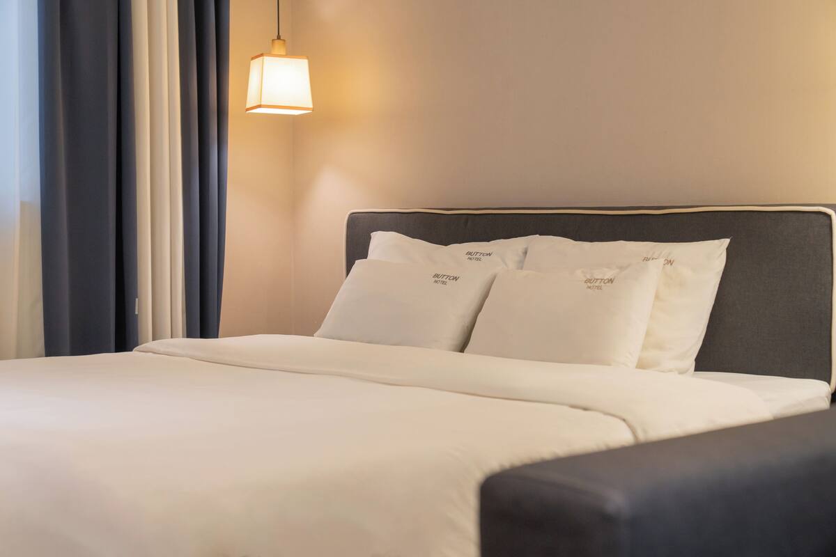 A hotel room with a neatly made bed, a headboard with pillows, and a hanging light fixture.