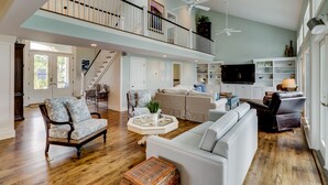 A spacious living room with a high ceiling, a staircase leading to an upper level, and a large flat-screen TV mounted on the wall.