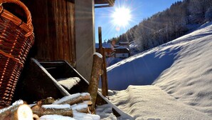 A wooden building with a basket and logs in the foreground, a snowy slope, and houses in the background. - Waldhütte - SKI in/SKI out - by "wachterhof" (Kaltenbach)
