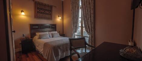 Une chambre avec un plancher en bois, un lit recouvert de draps blancs, une table de chevet, une chaise, un bureau et une fenêtre avec des rideaux.