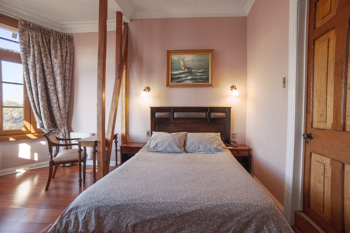 Un dormitorio con una cama grande, una mesita de noche, una silla, una puerta de madera y un cuadro de un velero en la pared.