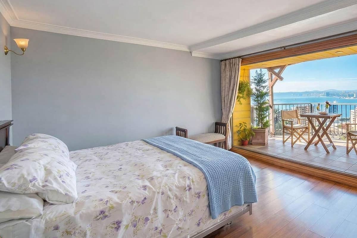 Un dormitorio con cama, mesita de noche, ventana con cortinas y vistas al exterior.