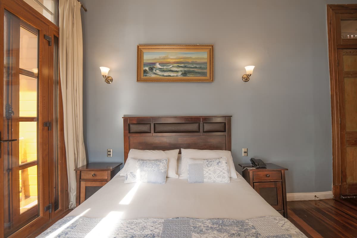 Una cama bien tendida con cabecera, dos mesitas de noche y un cuadro en la pared.
