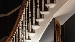 Un escalier avec des balustrades en bois et une main courante blanche, menant à un palier où se trouvent une lampe de table et un mur sombre.