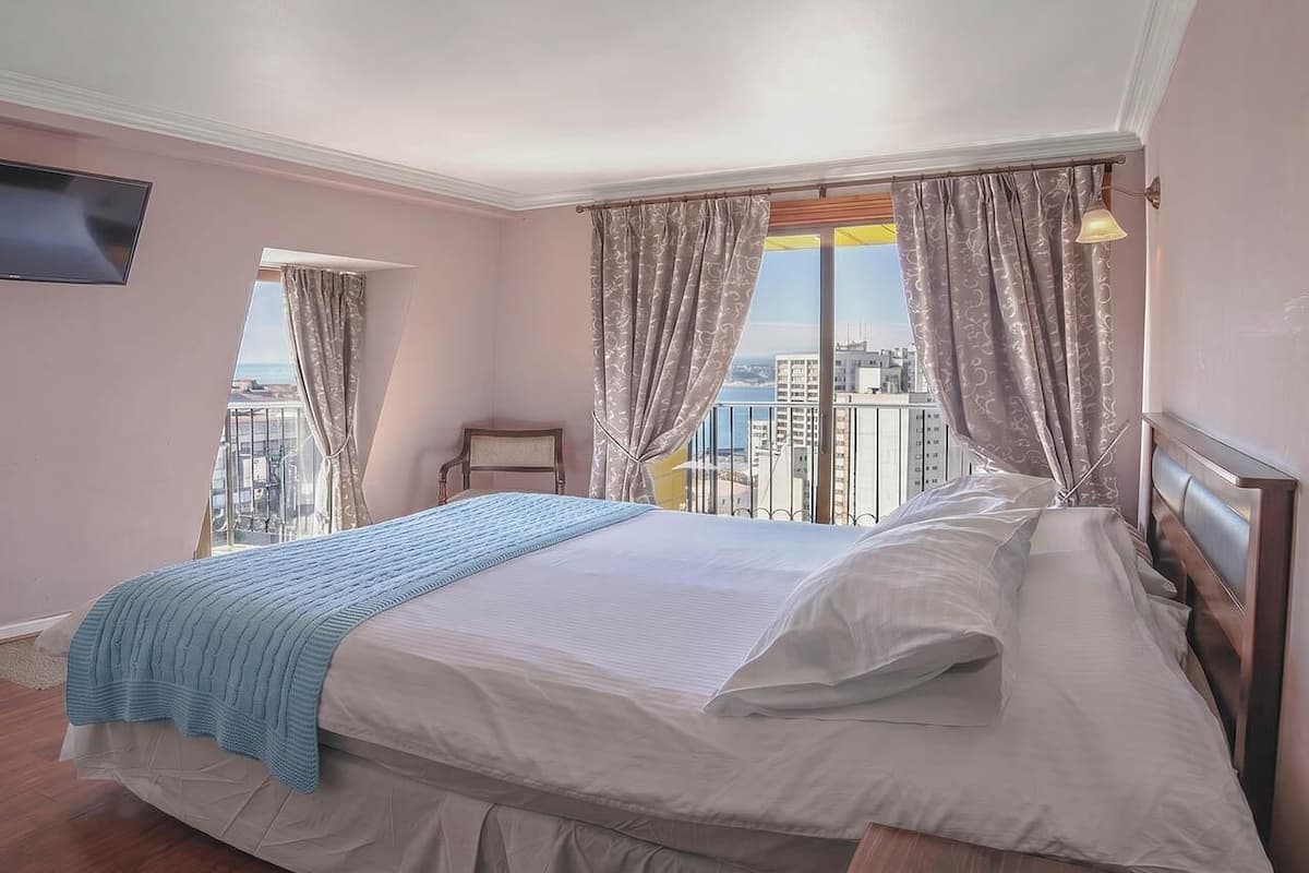 Un dormitorio con cama, cortinas, mesita de noche, lámpara y vistas a edificios.