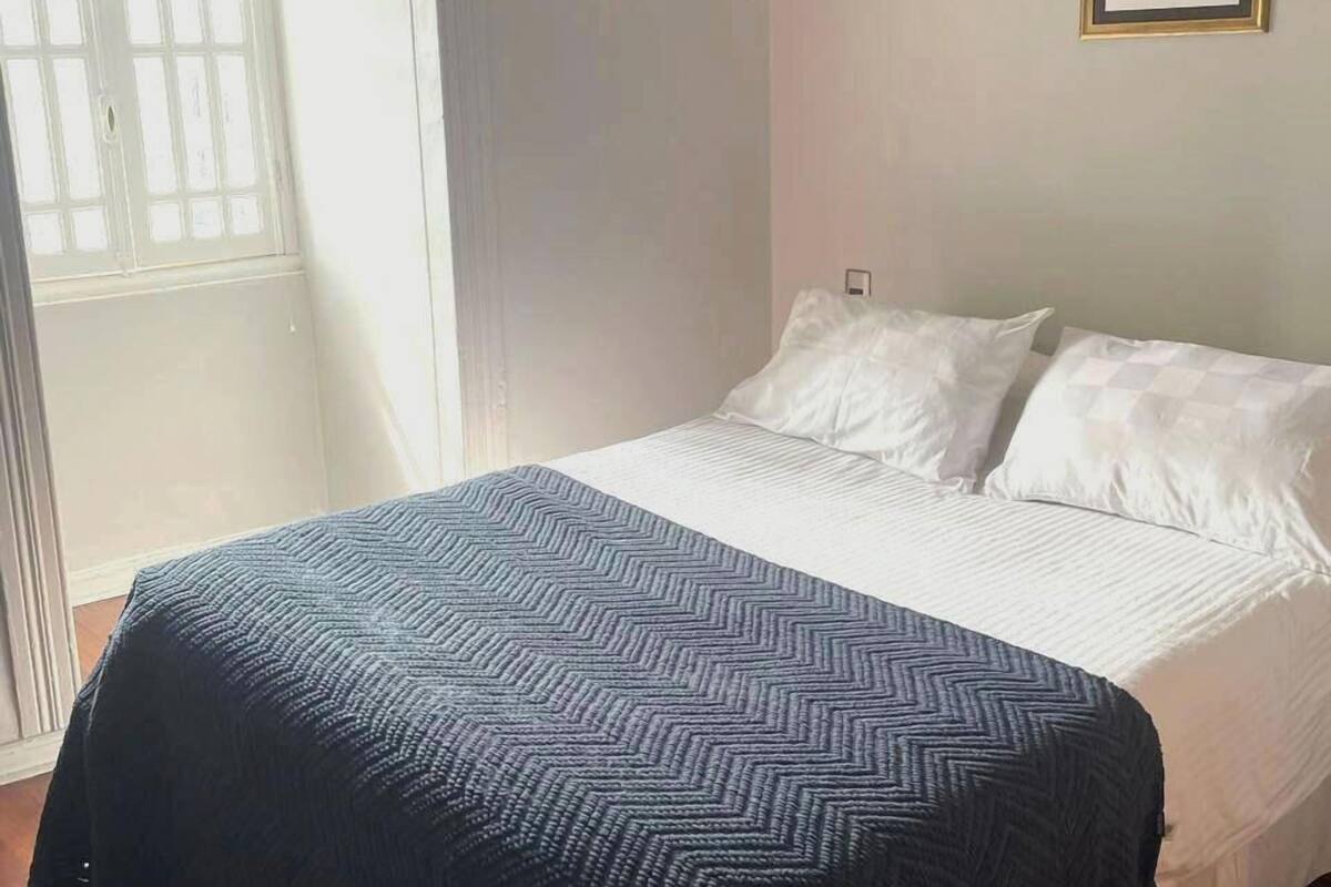 Un dormitorio con cama, ventana, lámpara de pared y un cuadro enmarcado.