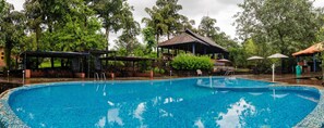 Una piscina con borde de azulejos, rodeada de árboles y estructuras cubiertas.